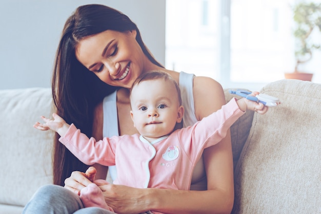 Hier bin ich! Schöne junge Frau, die Baby mit Spielzeug auf ihrem Kniesand hält und sie mit Liebe betrachtet, während sie zu Hause auf der Couch sitzt