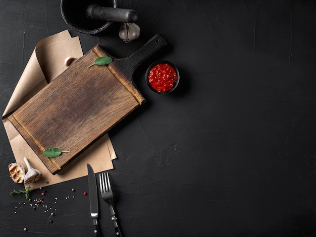 Hierbas en un tablero de madera pergamino tenedor y cuchillo ajo a la parrilla pimienta y salsa de sal mortero con mortero Fondo negro Primer plano Vista superior