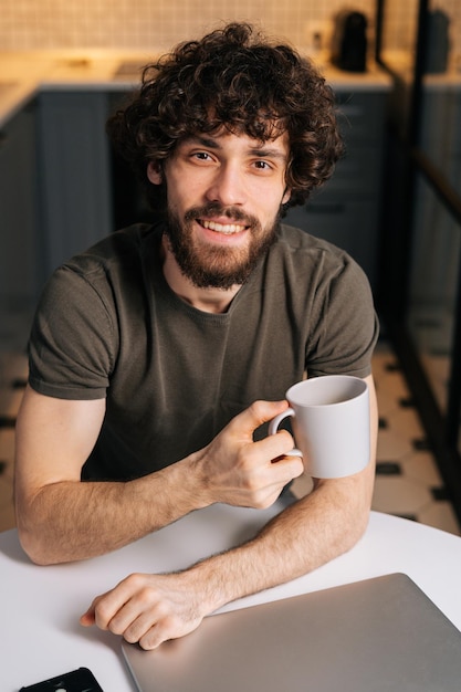 Highangle-Ansicht eines lächelnden bärtigen jungen Geschäftsmannes, der in der Hand Tasse mit Morgenkaffee hält, der mit geschlossenem Laptop-Computer am Tisch sitzt