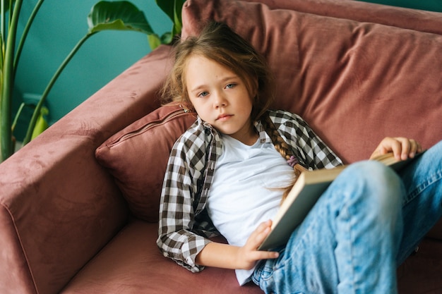 Highangle vista de infeliz adorable niño niña leyendo un libro de papel acostado en un sofá suave en casa