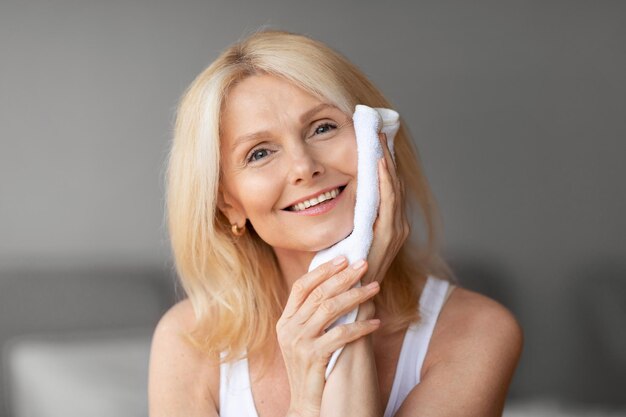 Higiene diaria hermosa mujer de mediana edad limpiando la cara con una toalla haciendo rutina matutina sonriendo