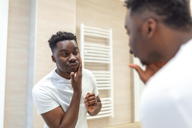 Higiene matinal Homem bonito no banheiro olhando no espelho Reflexo do homem africano com barba olhando no espelho e tocando o rosto no banho