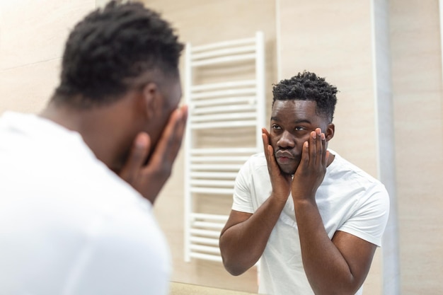 Higiene matinal Homem bonito no banheiro olhando no espelho Reflexo do homem africano com barba olhando no espelho e tocando o rosto no banho