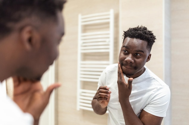 Higiene matinal Homem bonito no banheiro olhando no espelho Reflexo do homem africano com barba olhando no espelho e tocando o rosto no banho