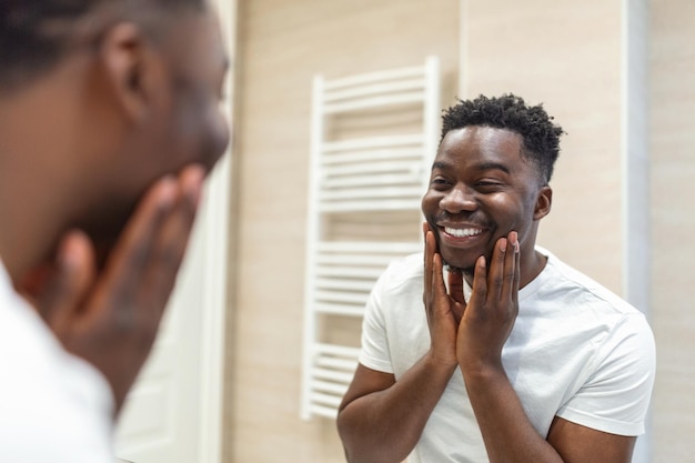 Higiene matinal homem bonito no banheiro olhando no espelho reflexo do homem africano com barba olhando no espelho e tocando o rosto no banho