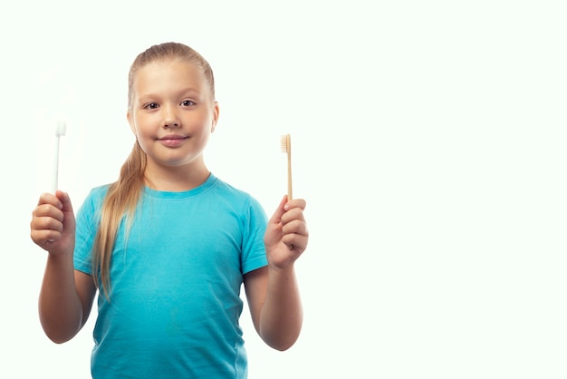 Higiene oral. caucasiana menina bonitinha segurando escovas de dentes de bambu e plástico nas mãos. fundo branco, lugar para texto. conceito de seleção de produtos ecológicos.