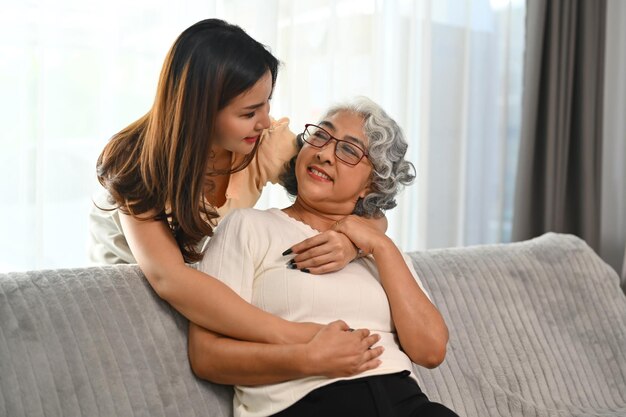 Foto hija adulta sonriente parada detrás del sofá y abrazando a la madre mayor concepto de vinculación familiar