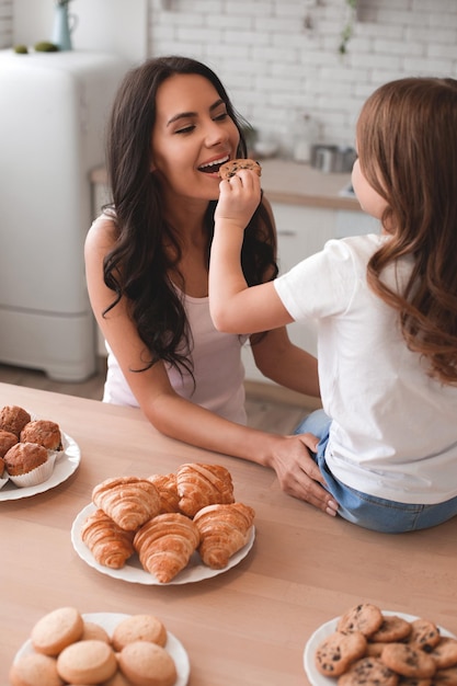 Hija alimentando a la madre con una galleta sentada en la mesa de la cocina