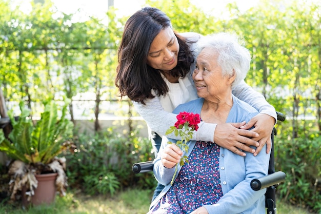 La hija del cuidador abraza y ayuda a una anciana o anciana asiática sosteniendo una rosa roja en silla de ruedas en el parque