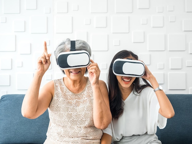 La hija feliz de una joven asiática y su madre, una mujer mayor que usa gafas VR, disfrutan de un juego 3d en línea juntos en una habitación blanca Mamá e hija con el concepto de estilo de vida de tecnología virtual de realidad