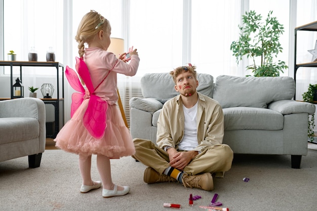 Hija haciendo una foto de su padre exhausto con maquillaje y peinado divertidos, pasatiempo divertido en casa con sus padres