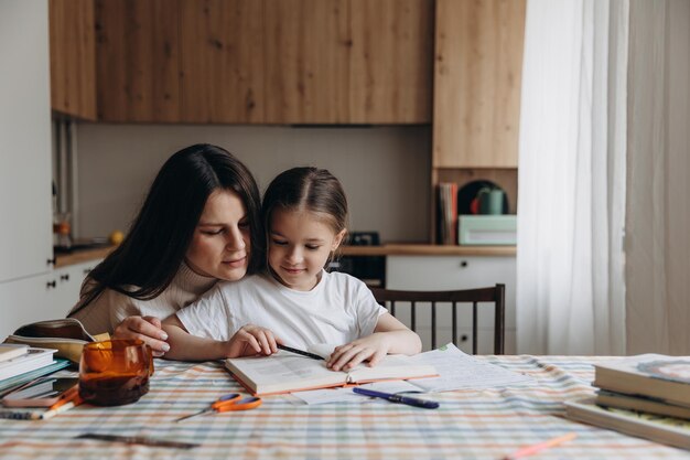 Foto hija haciendo la tarea junto con la madre joven en casa en la mesa de la cocina