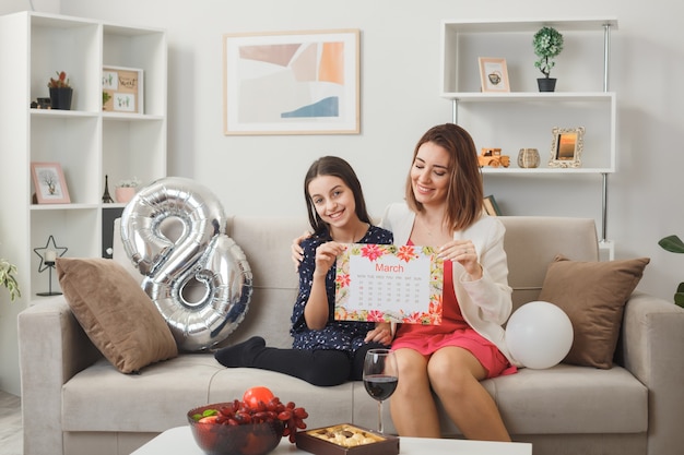 Hija y madre sonriente en el día de la mujer feliz con postal sentado en el sofá en la sala de estar