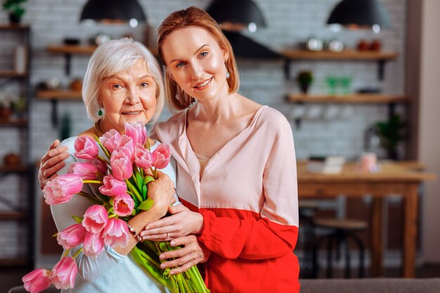 Hija y madre con tulipanes. Hija madura pelirroja atractiva en blusa rosa posando con el envejecimiento de la madre de pelo plateado sonriente buena atractiva con ramo de tulipanes