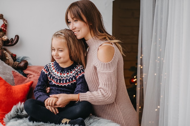 Hija con mamá en la ventana esperando la Navidad. El tierno abrazo de la madre. Comunicación afectuosa entre mamá e hija.