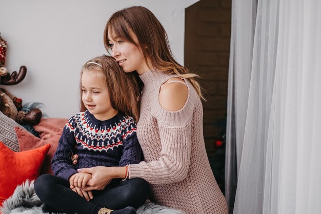 Hija con mamá en la ventana esperando la Navidad. El tierno abrazo de la madre. Comunicación afectuosa entre mamá e hija.