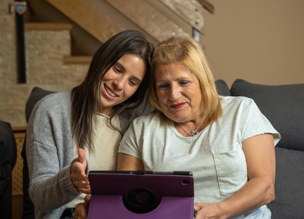 La hija le muestra a su madre algo en la tableta y la madre está atenta mientras están felizmente sentadas en el sofá de casa