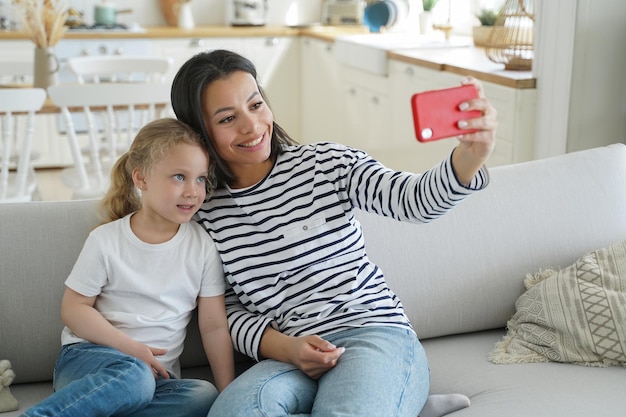 La hija pequeña de la mamá feliz se toma una selfie o hace una videollamada sosteniendo el teléfono sentado en el sofá