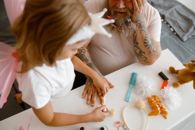 Hija pinta las uñas de papá con esmalte amarillo brillante en una mesa blanca en la habitación