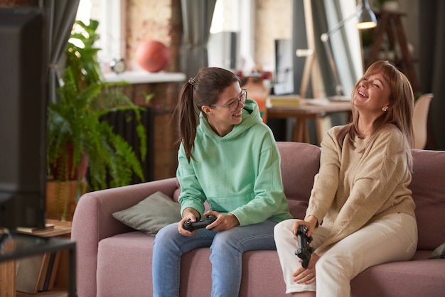 Hija riendo junto con su madre en el sofá mientras juegan videojuegos en casa