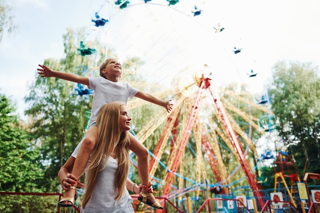 La hija se sienta sobre los hombros. Niña alegre que su madre pasa un buen rato en el parque junto a las atracciones.