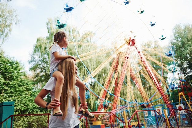 La hija se sienta sobre los hombros. Niña alegre que su madre pasa un buen rato en el parque junto a las atracciones.