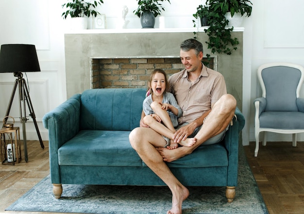 Hija sonriente sentada sobre las rodillas del padre en el sofá