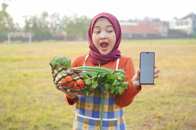 Hijab mujer sosteniendo verduras en una cesta que muestra el teléfono en blanco