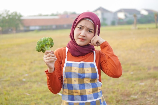 Hijab mujer usar delantal sostener brócoli y gesto pensar algo