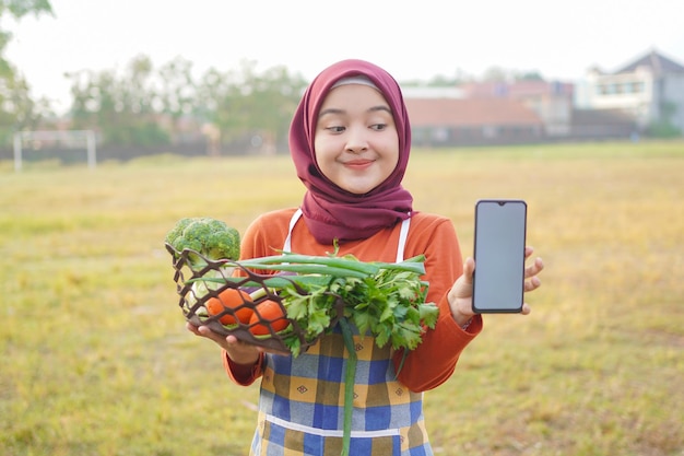 Foto hijab mulher segurando legumes em uma cesta mostrando telefone em branco