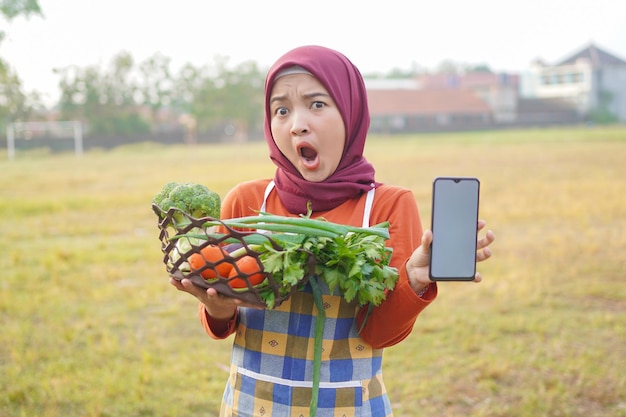 Hijab mulher segurando legumes em uma cesta mostrando telefone em branco