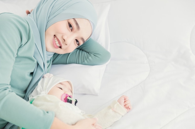 Hijab musulmán quedarse con su bebé en la cama blanca sonriendo en casa, cuidado de la madre infantil.