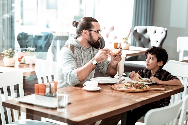 Foto hijo encantador. radiante hombre barbudo con gafas mirando a su encantador hijo mientras come pizza para el almuerzo