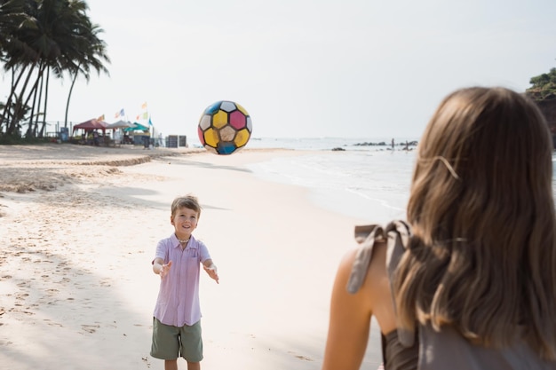 Hijo feliz jugando a la pelota con mamá en la playa en un día soleado