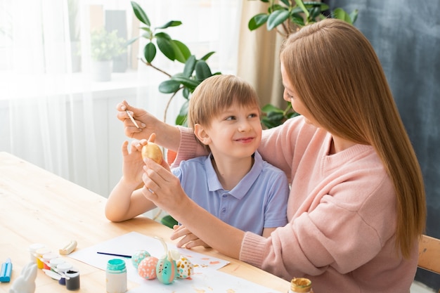 Hijo sonriente mirando a la madre mientras ella pinta el huevo de Pascua en su mano en casa