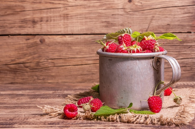 Himbeeren-Hintergrund. Reife Früchte in einem Vintage-Becher im rustikalen Stil. Modernes hartes Licht, dunkler Schatten. Alter Holzbretttisch, Kopierraum