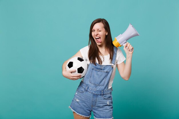 Una hinchada de fútbol joven y divertida apoya al equipo favorito con un megáfono de balón de fútbol que muestra la lengua aislada en un fondo azul turquesa. Emociones de la gente, concepto de ocio familiar deportivo. Simulacros de espacio de copia.