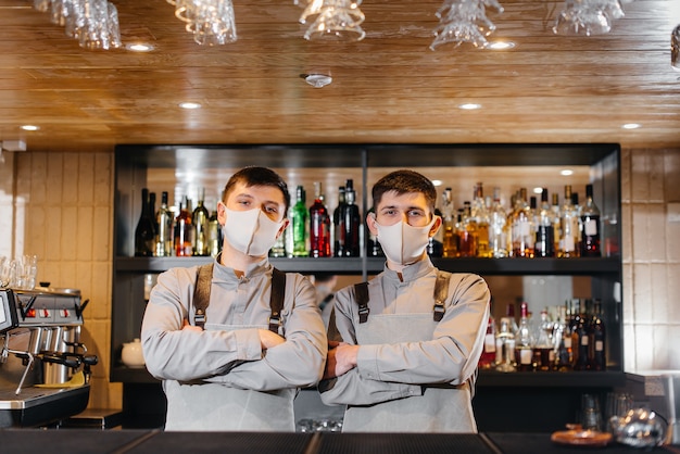 Hinter der Bar stehen zwei stilvolle Barkeeper in Masken und Uniformen während der Pandemie. Die Arbeit von Restaurants und Cafés während der Pandemie.