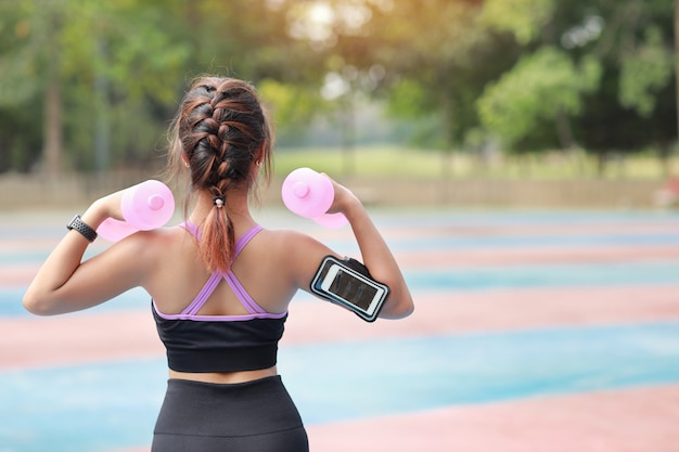 Hintere Ansicht athletische junge Schönheit asiatische Frau in Sportbekleidung stehend und Hanteln im Freien für Morgenübung heben. Aktives Mädchen trainieren auf Bizepsübung mit grünem Baumhintergrund. Sportkonzept