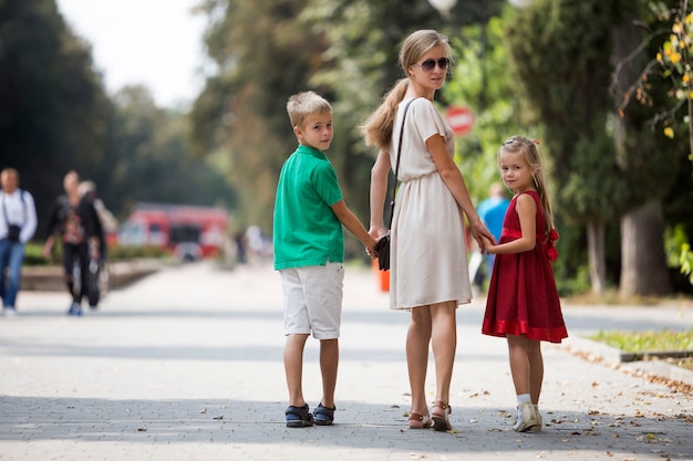 Hintere Ansicht der glücklichen Familie, junge blonde langhaarige Frau, die zurück Händchenhalten mit zwei netten Kindern, Tochter und Sohn auf sonniger Parkgasse auf grünem Bäume bokeh am warmen Sommertag schaut.