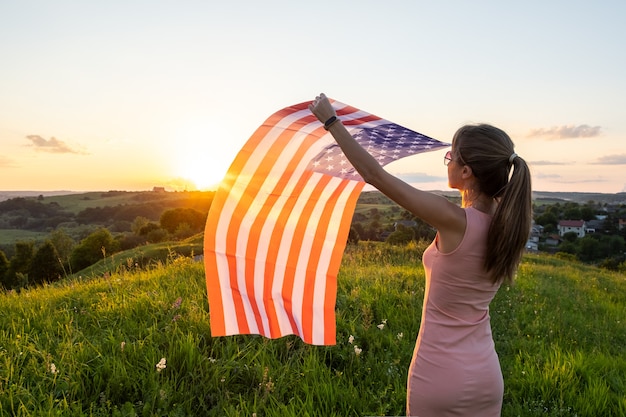 Hintere Ansicht der glücklichen Frau mit USA-Nationalflagge, die draußen bei Sonnenuntergang steht. Positive Frau, die den Unabhängigkeitstag der Vereinigten Staaten feiert. Konzept des Internationalen Tages der Demokratie.