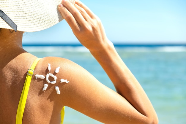 Hintere Ansicht der jungen Frau, die am Strand mit Sonnenschutzcreme in der Sonnenform auf ihrer Schulter bräunt. UV-Sonnenbrandschutz und Sonnenschutz-Hautpflegekonzept