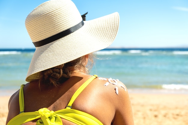 Hintere Ansicht der jungen Frau, die am Strand mit Sonnenschutzcreme in der Sonnenform auf ihrer Schulter bräunt