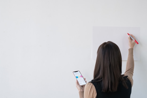 Hintere Ansicht der jungen Geschäftsfrau oder des Lehrers mit Smartphone und Textmarker, die durch Whiteboard stehen