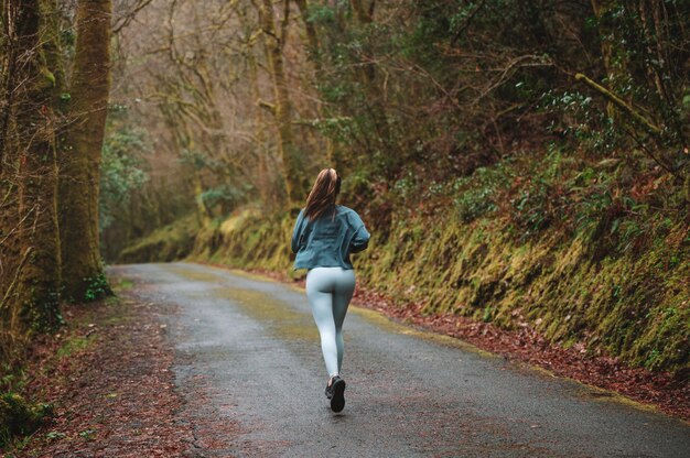 Hintere Ansicht der nicht erkennbaren Sportlerin in Sportbekleidung, die entlang der Asphaltstraße in Wäldern während des Cardio-Trainings läuft