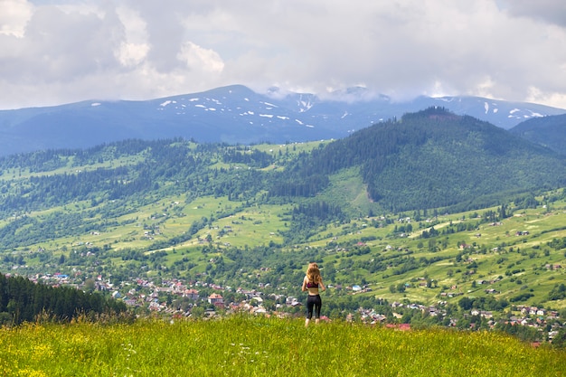 Hintere Ansicht der schlanken jungen Frau, die auf grasartigem Tal, grünen Bergen am sonnigen Sommertag steht