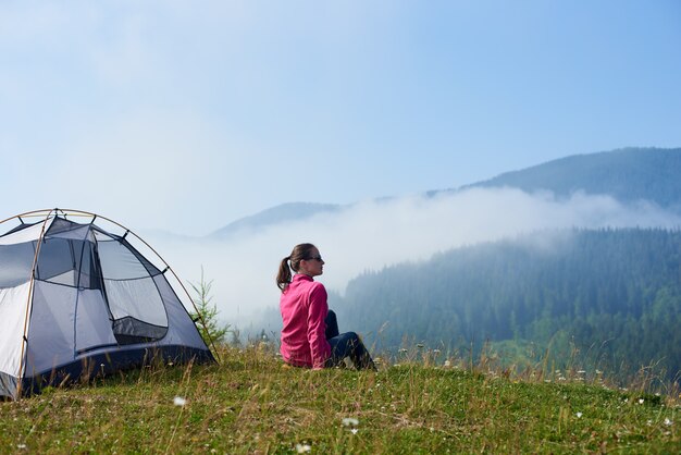 Hintere Ansicht des jungen Touristenmädchens, das auf grünem Gras des blühenden Tals sitzt