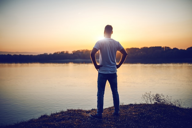 Hintere Ansicht des schönen kaukasischen Mannes, der mit den Händen auf den Hüften auf der Klippe steht und Fluss und schönen Sonnenuntergang betrachtet.