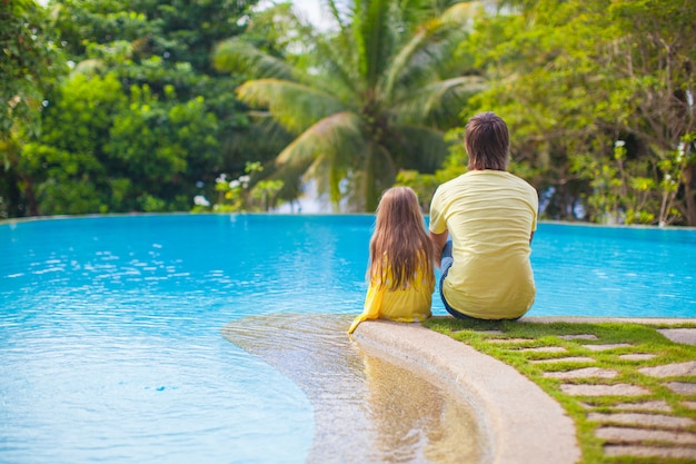 Hintere Ansicht eines jungen Vaters und seiner netten Tochter, die durch den Swimmingpool sitzen