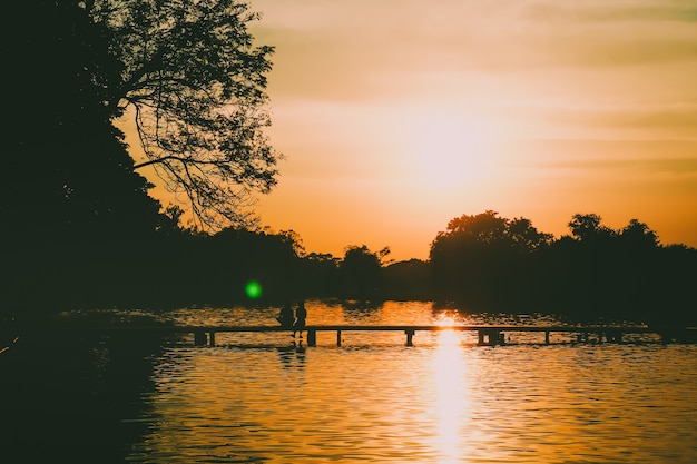 Hinteres Schattenbild eines Paares auf dem schönen Waldsee der Brücke am Sonnenunterganghintergrund